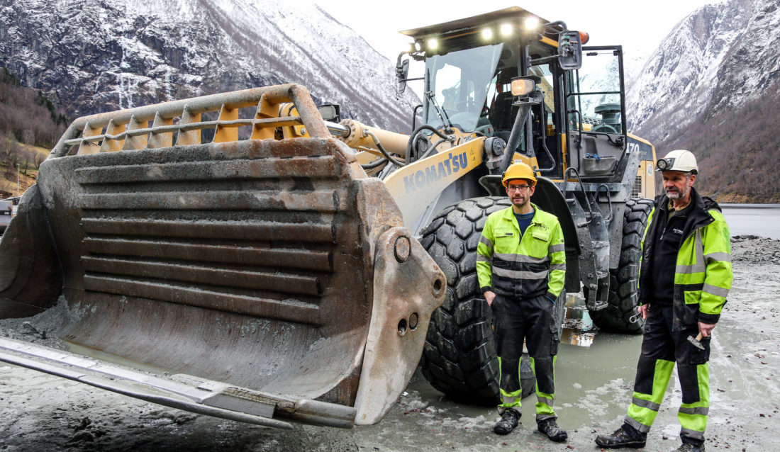 Far og sønn, Jan Arild (til høyre) og Atle Bjordal, driver A og J Bjordal AS, som er involvert i en kjempestor kraftverkutbygging. Her ved siden av nyvinningen deres: En klaff som festes på den åpne siden av sidetippskuffen, som kan festes i to posisjoner. Når den er framme/oppe, fungerer den som en fast side og skuffen kan brukes til lasting, mens når den en bakover/nede (som her) kan man tippe til siden på vanlig måte. (Foto: Runar F. Daler).